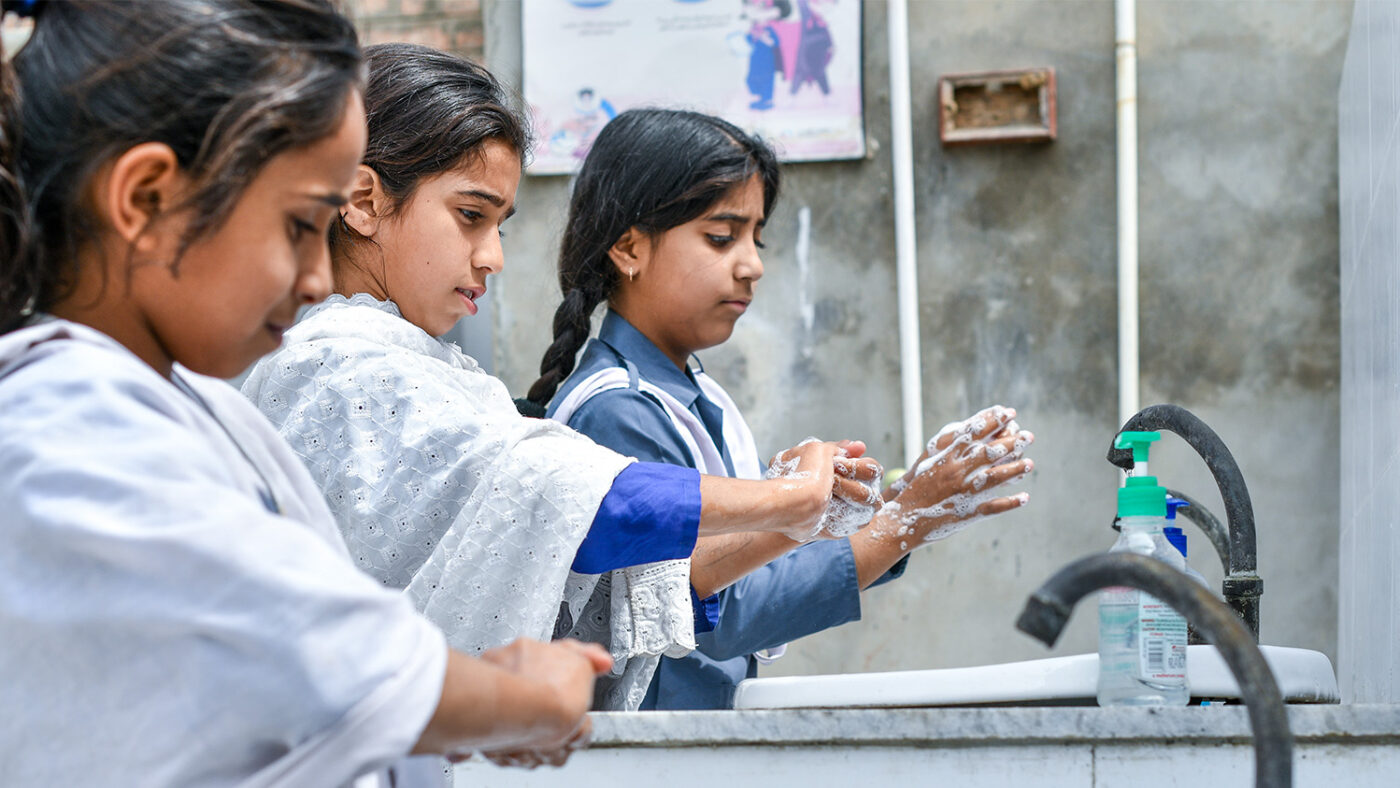 Three children wash their hands with soap at a sink.