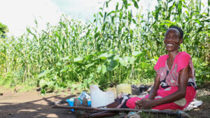 Eunice sits on the ground next to field where she's warming sorghum. A pair of crutches are lying next to her.