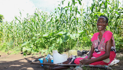 Eunice sits on the ground next to field where she's warming sorghum. A pair of crutches are lying next to her.