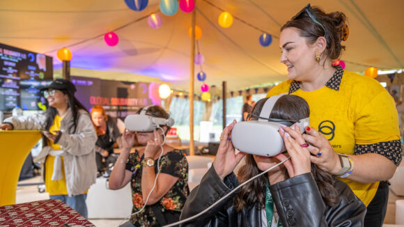 Two women wear virtual reality headsets at an event in Ireland. One of the women is supported by another women wearing a Sightsavers t-shirt.