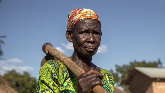 Sofia stands outside her garden in Uganda, holding a mattock over her right shoulder.