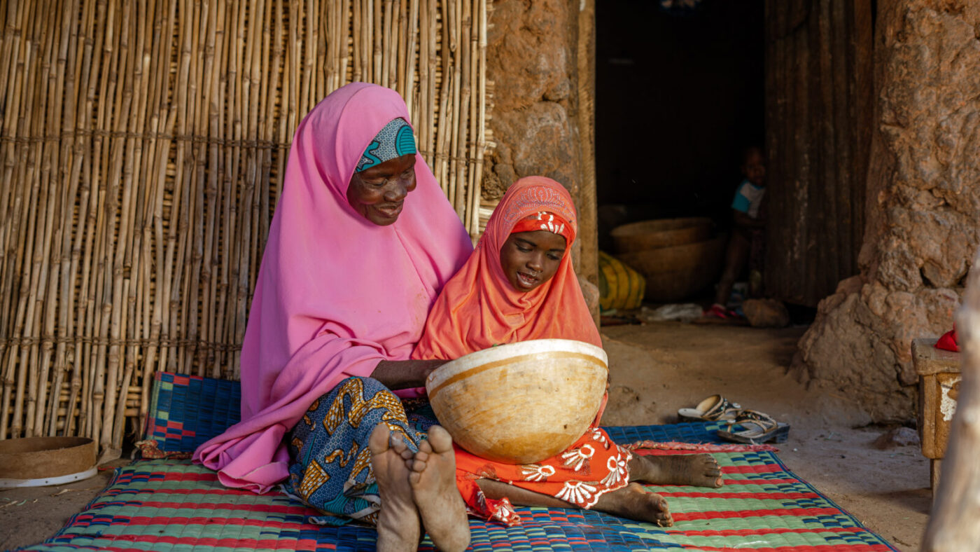 Aisha sits on the floor with a family member. They're both smiling and looking inside a basket.