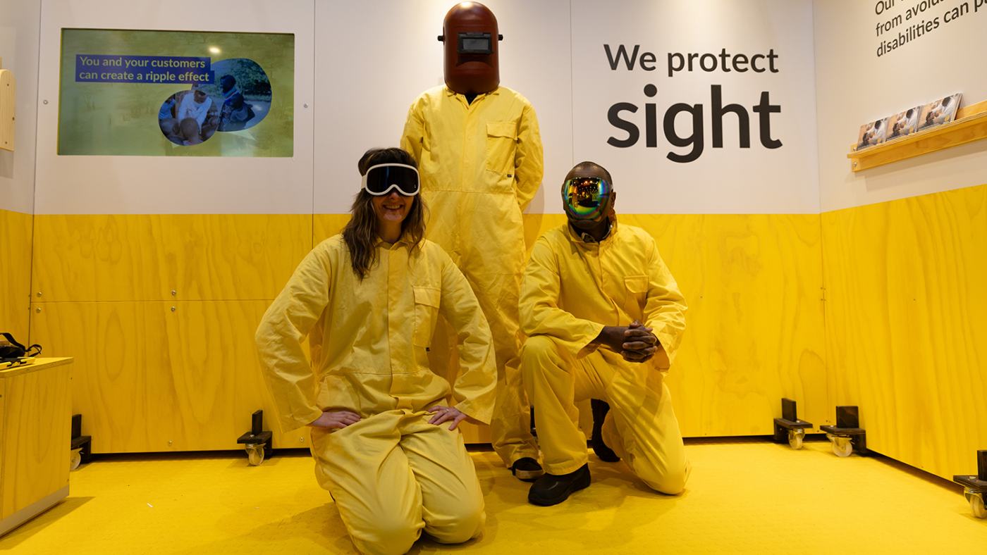 Sightsavers staff wear yellow boiler suits and protective eyewear as they pose for a picture in front of the Sightsavers stand.