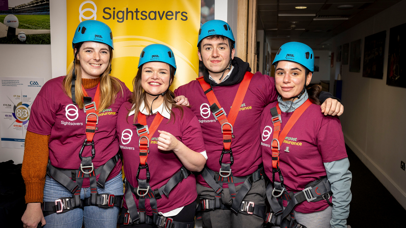 Participants smile wearing helmets and harnesses with a Sightsavers poster in the background.