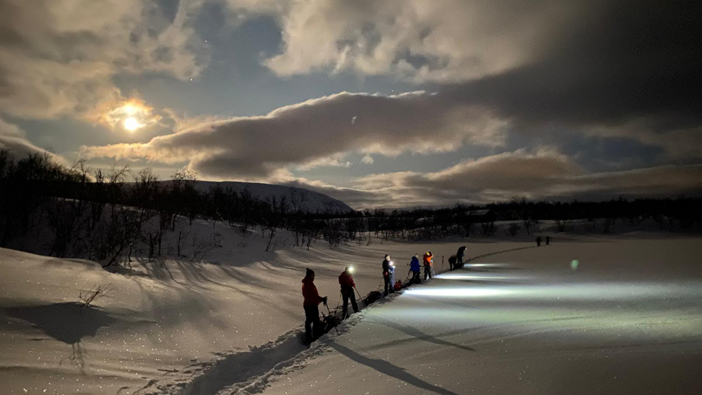 The team travels across a snowy Arctic landscape using skis and pulling a bag of supplies behind them.