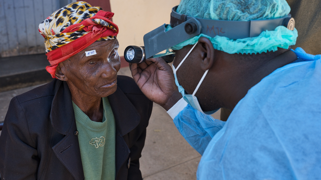 Belita, an older woman, has her eyes checked for trachoma with a torch by Dr Mwbeku.