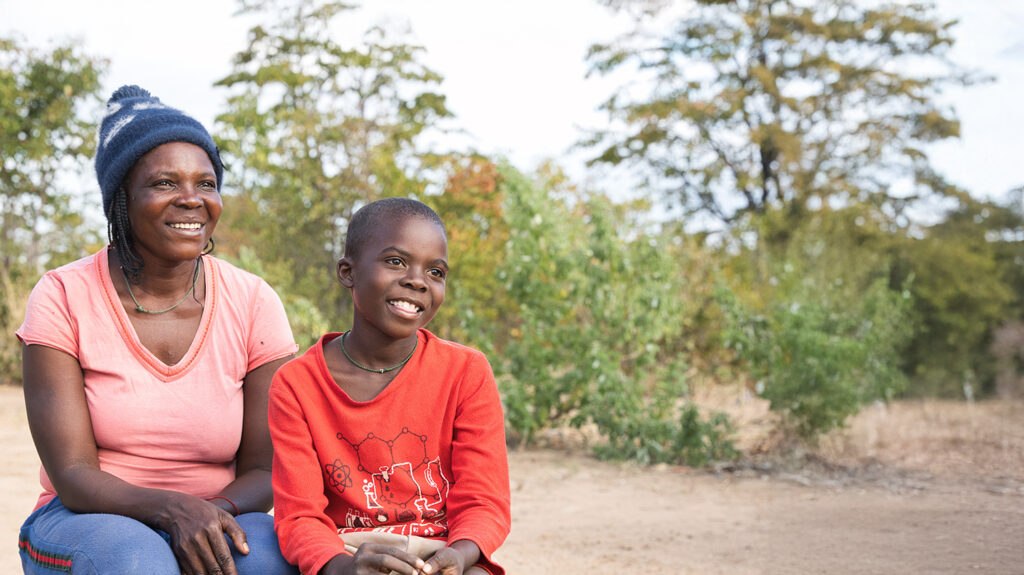 Enitah and her son Ariel sit outside, near their home in Zimbabwe. They're both smiling in the sunshine. The ground is sandy, with trees and bushes in the background.