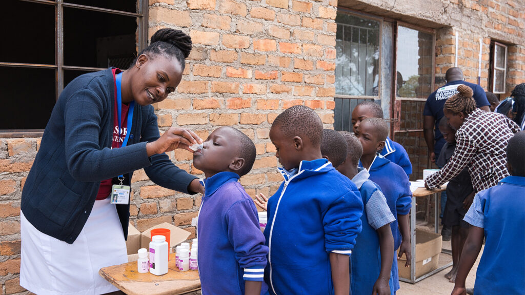 In Zimbabwe, health worker Melody gives a boy a cup of liquid to drink during a mass drug administration campaign at his school. Other children stand behind him in the line, wearing school uniform.