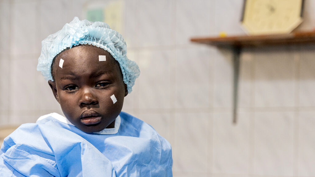 Six-year-old Happy from Uganda sits in hospital after eye surgery. She's wearing surgical scrubs and a hat, with surgical stickers on her face.