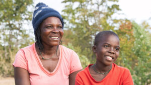 Enitah and her son Ariel sit outside near their home in Zimbabwe. They're both smiling in the sunshine. The ground is sandy, with trees and bushes in the background.