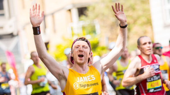 James cheers while running the 2025 London Marathon. He's wearing a Sightsavers running vest with his name on the front.