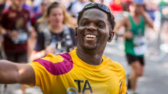 Fundraisier Taofeek waves while running the 2025 London Marathon. He's wearing a yellow Sightsavers T-shirt.