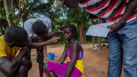 A man examines a young girl's eye. There are trees in the background.