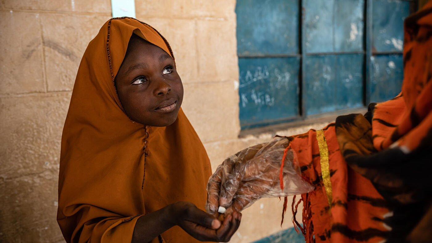 A girl wearing a hijab is handed a white tablet to protect her from intestinal worms.