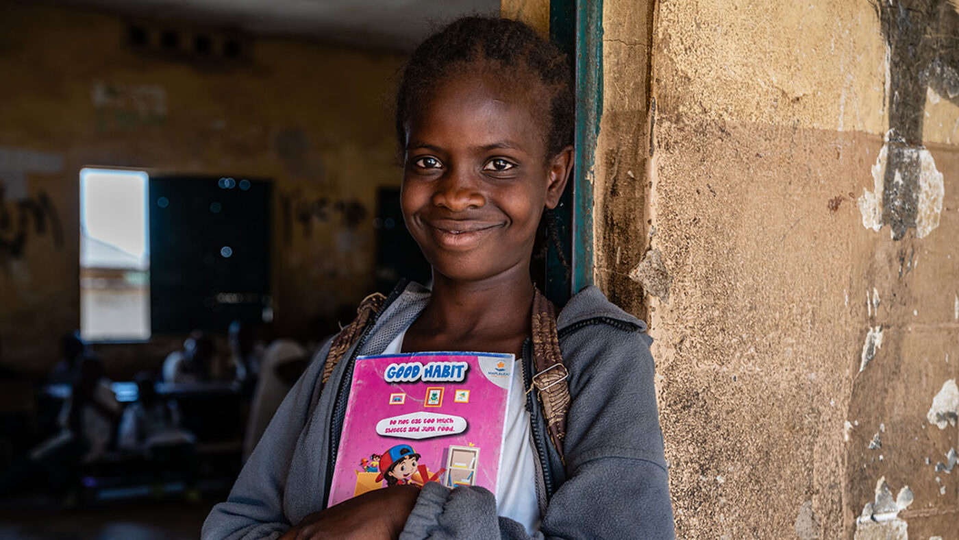 Favor leans against a wall with book in her arms outside her classroom at school.