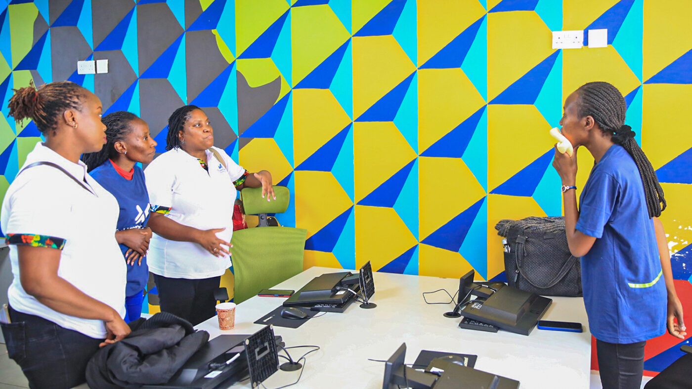Maryanne stands over a laptop at a desk. Three women from Standard Chartered watch from the other side of the desk.