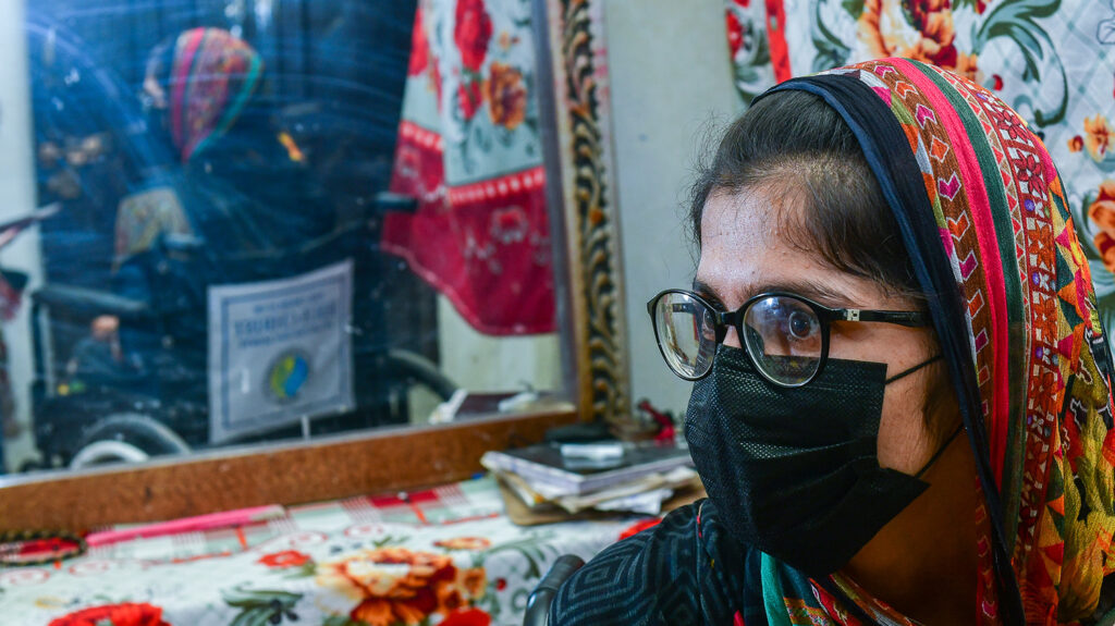 Aasiya sits next to a mirror wearing her glasses and a medical mask.