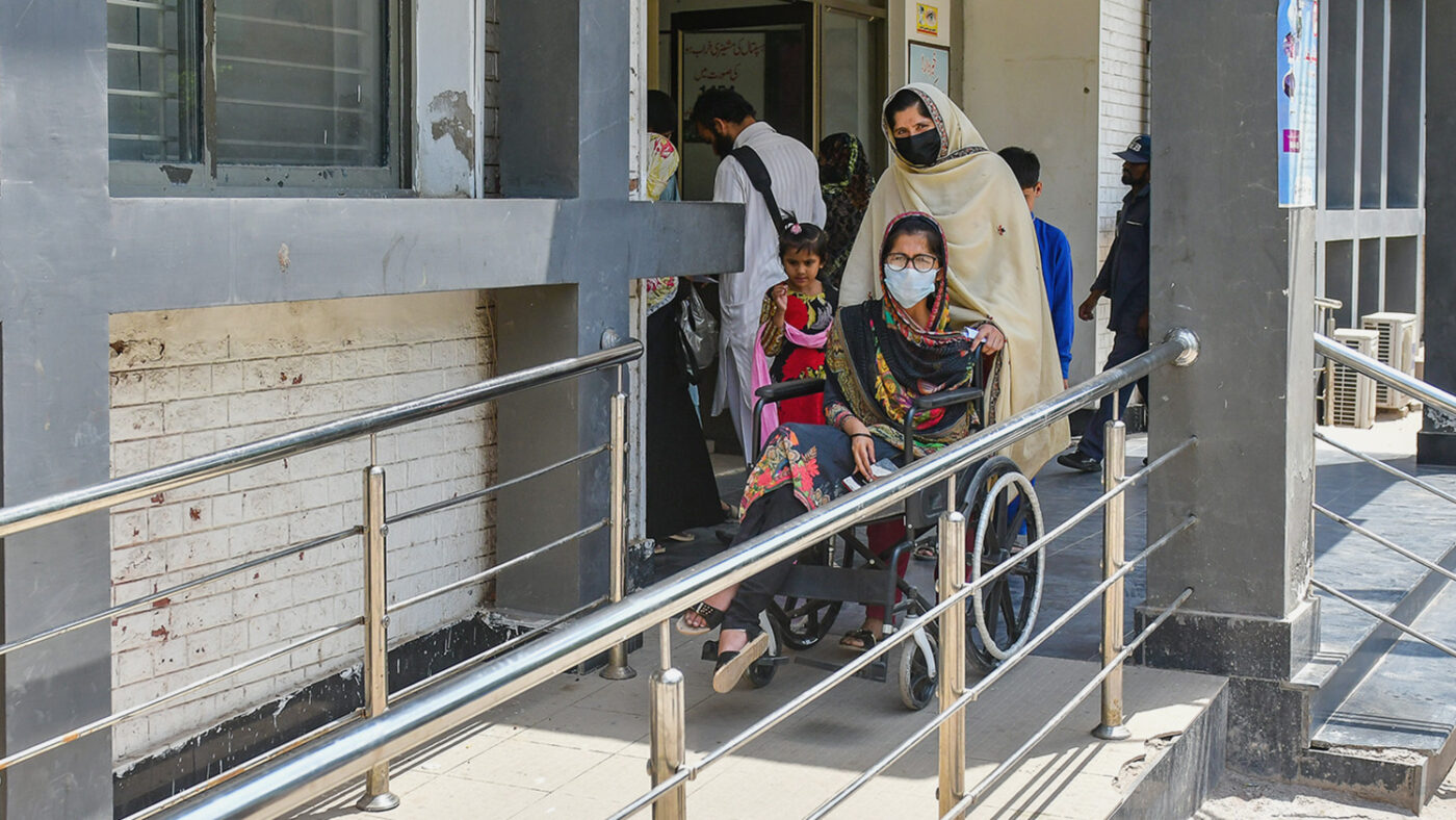 Aasiya's sister-in-law pushes Aasiya's wheelchair down a ramp outside a building. They are both wearing medical masks.