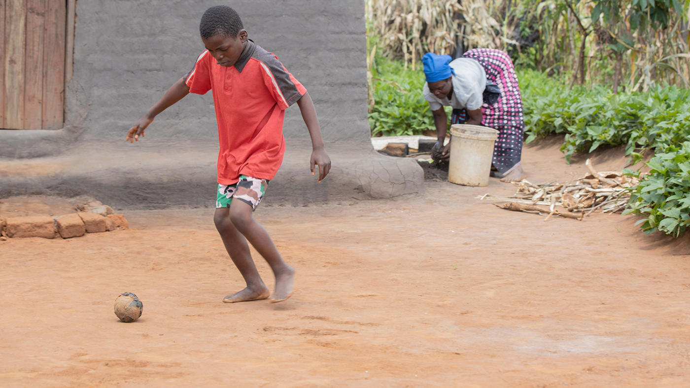 Chisomo kicks a homemade football in front of his home. His mother Margaret is working in the background.