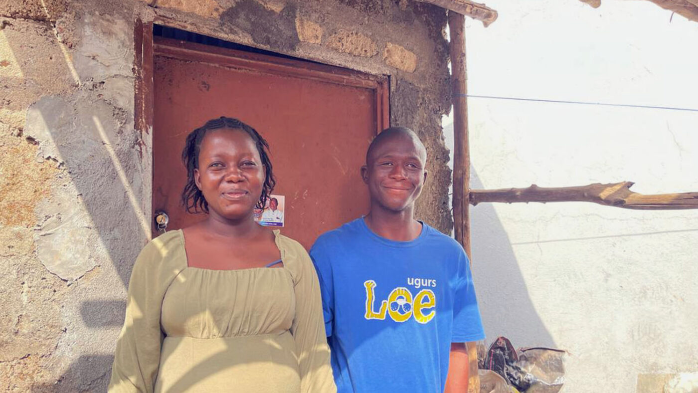 Magdalene and Abubakarr stand in the shade outside a building. They're both smiling.