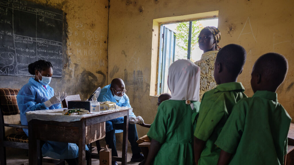 In a classroom, children queue to give blood samples. Two health workers wearing medical scrubs and masks are sitting at a desk. The child at the front is holding out their finger.