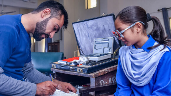 A male eye health worker conducts an eye test for a schoolgirl. She is wearing optometry glasses.