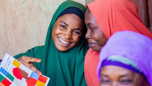 Two women wearing hijabs smile during a workshop. One is holding an illustrated booklet about family planning.