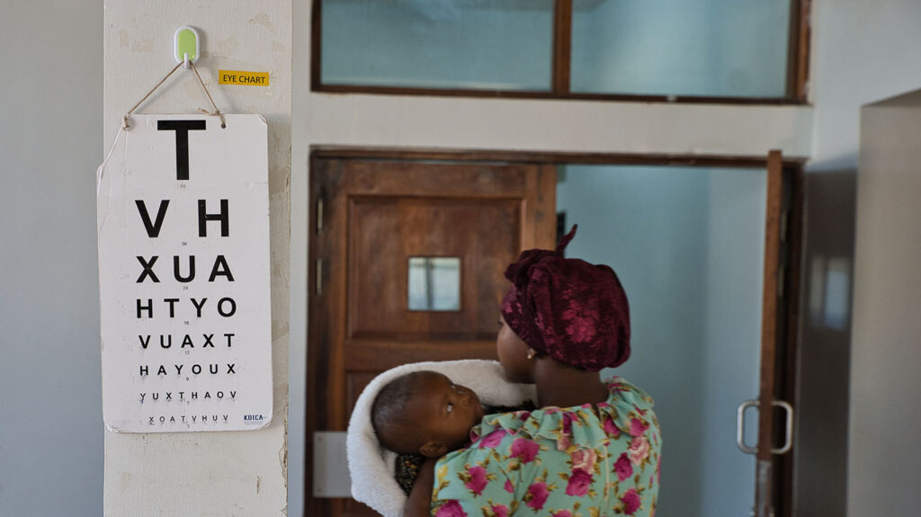A woman holds a small child, who is wrapped in a blanket. The woman is looking at an eye test chart on a wall in a hospital.