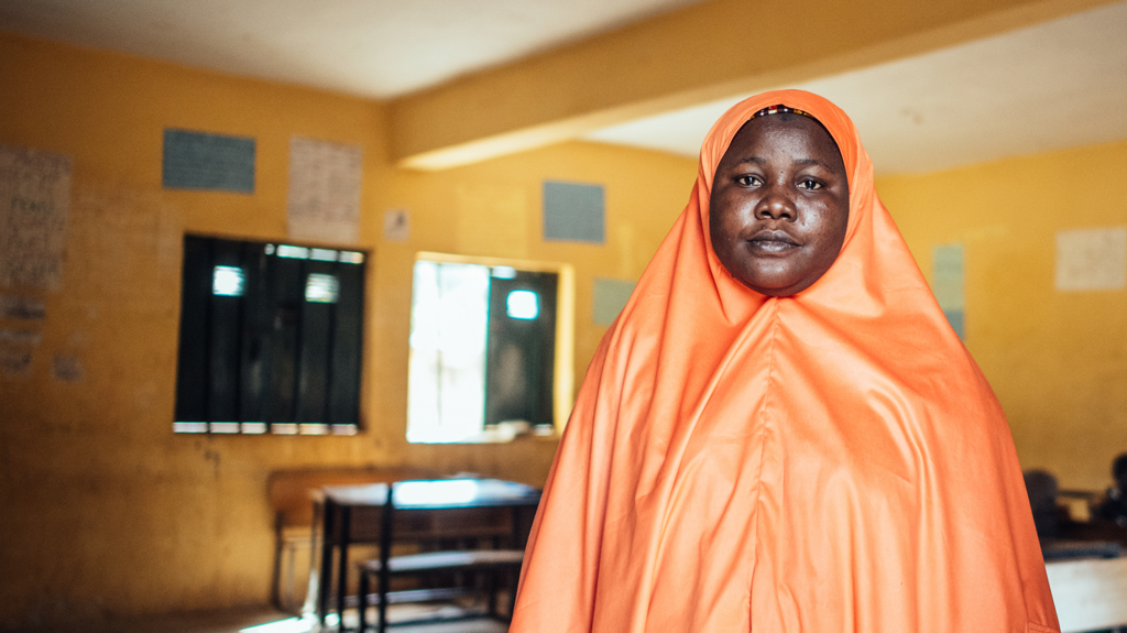 A woman stares into the camera. She wears an orange floor-length gown and khimar.