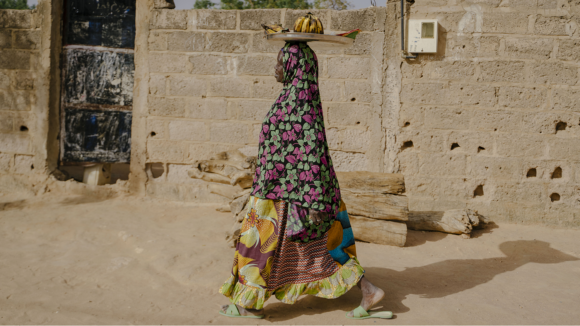 Une femme vêtue de vêtements traditionnels africains se promène dans une rue tout en équilibrant un plateau de fruits sur sa tête.