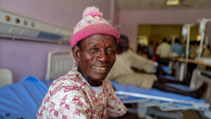 A man wearing a beanie hat smiles while sitting on a hospital bed.