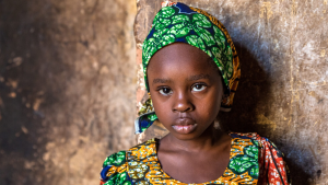 A young girl leans on a wall with a neutral expression on her face.