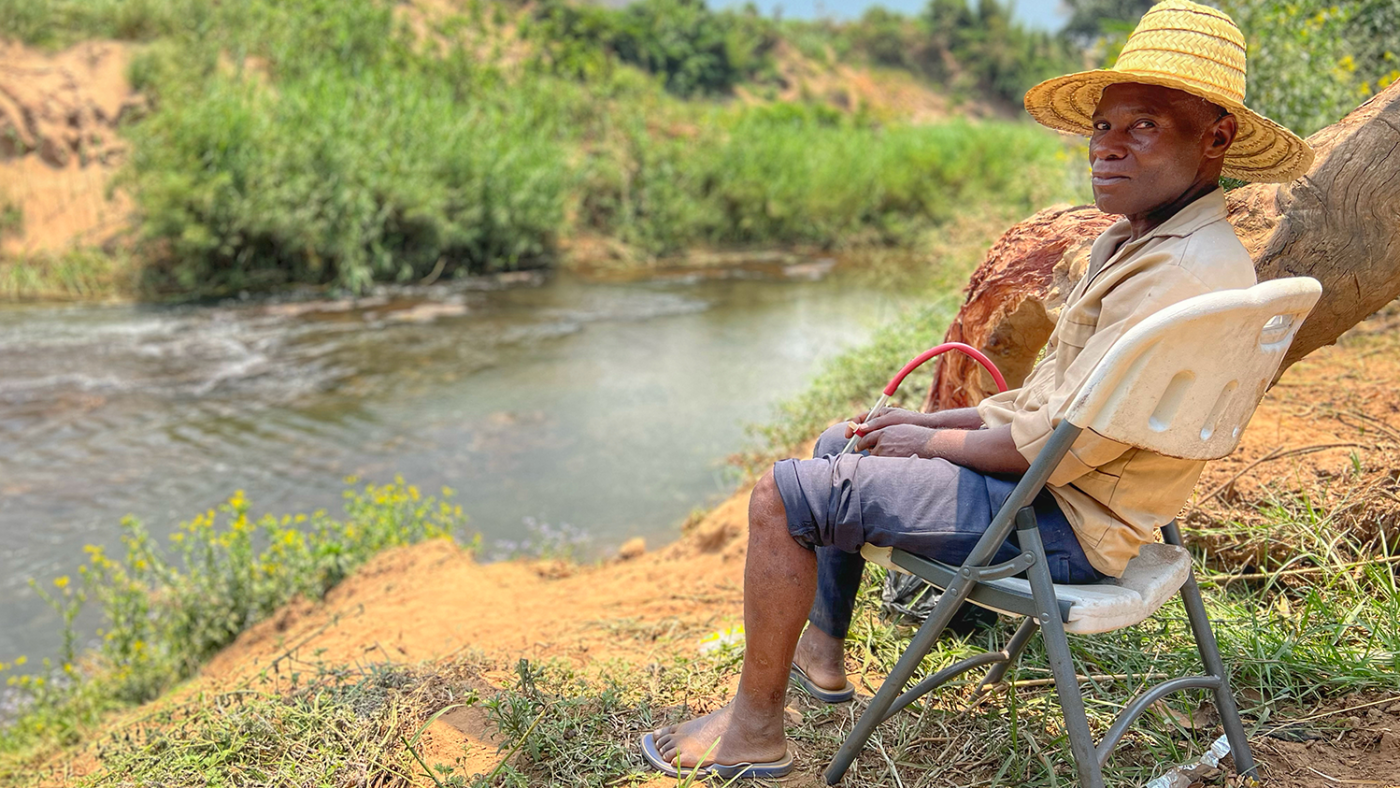 A man wearing a straw hat sits on a chair by a river.