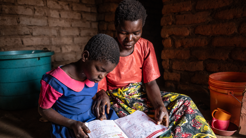 Melia, young girl reading from a school book, sat next to her mother, Veronica, in a brick house.