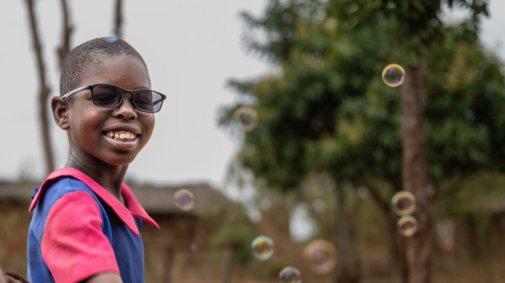 A girl smiles while wearing dark glasses. Bubbles float in the air around her.