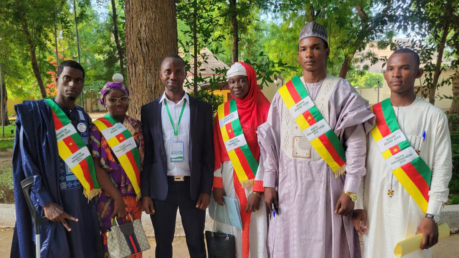 A group of men and women stand together outside. All of them are wearing colourful sashes in the colours of Cameroon, except for one man, who is wearing a suit.