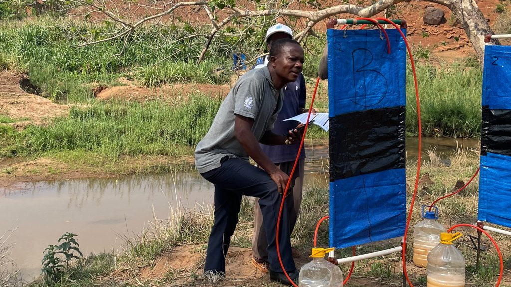 Two men standing by a river stare at a black fly trap, which consists of a square piece of blue tarpaulin attached to a wooden frame. There are also tubes that lead into water containers.