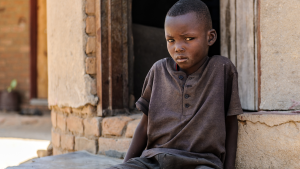 A young boy, Bretty, looks at the camera while sat on the ground outside a doorway.