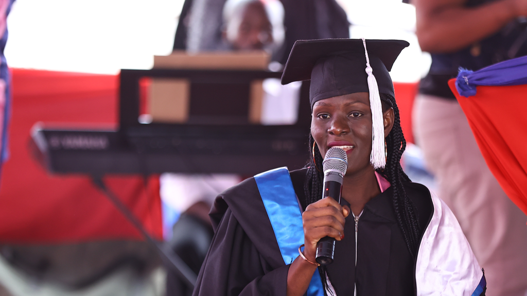 A woman wearing graduation attire speaks into a microphone while onstage.