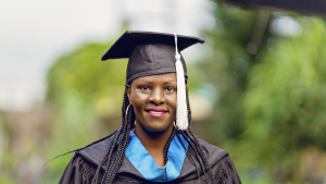 A woman wearing a mortarboard and a graduation robe with a blue sash. She has long braids and gold hoop earrings.