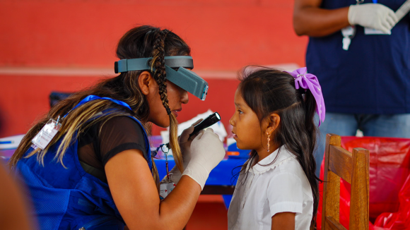 A woman wearing medical examination glasses shines a torch into a young girl's eyes to check for trachoma.
