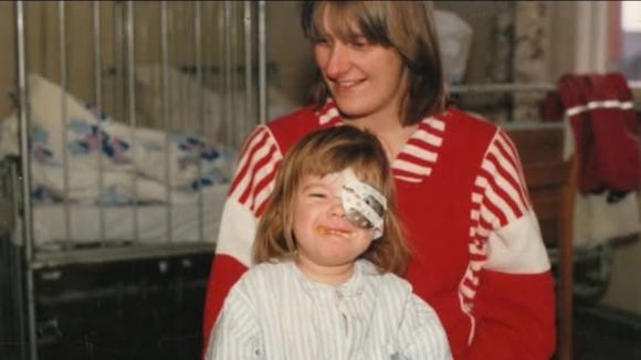 A toddler sits on her mother's lap in a hospital setting. The young girl is wearing a protective medical eye shield over her left eye.