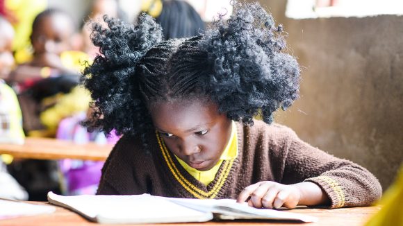 A girl with her hair in pigtails tilts her head towards an open book. She is sitting at a desk in a classroom.