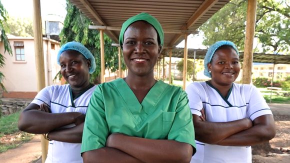 Dr Gladys Atto, wearing surgical scrubs, stands with two of her colleagues outside a hospital in Uganda. They have their arms crossed and are smiling at the camera.