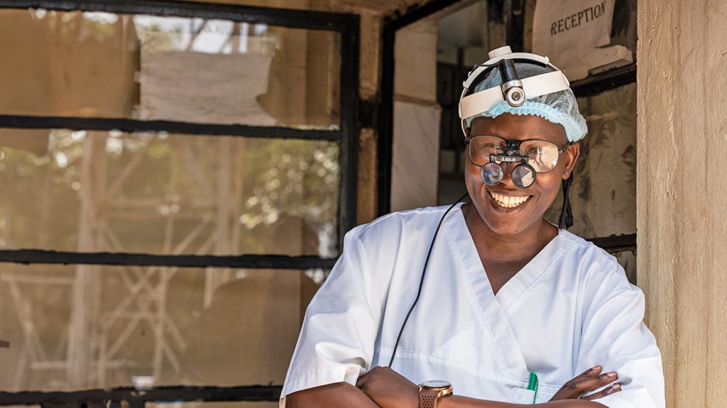 A trachoma surgeon wearing surgical scrubs and optical headwear smiles broadly and crosses his arms outside a clinic in Uganda.