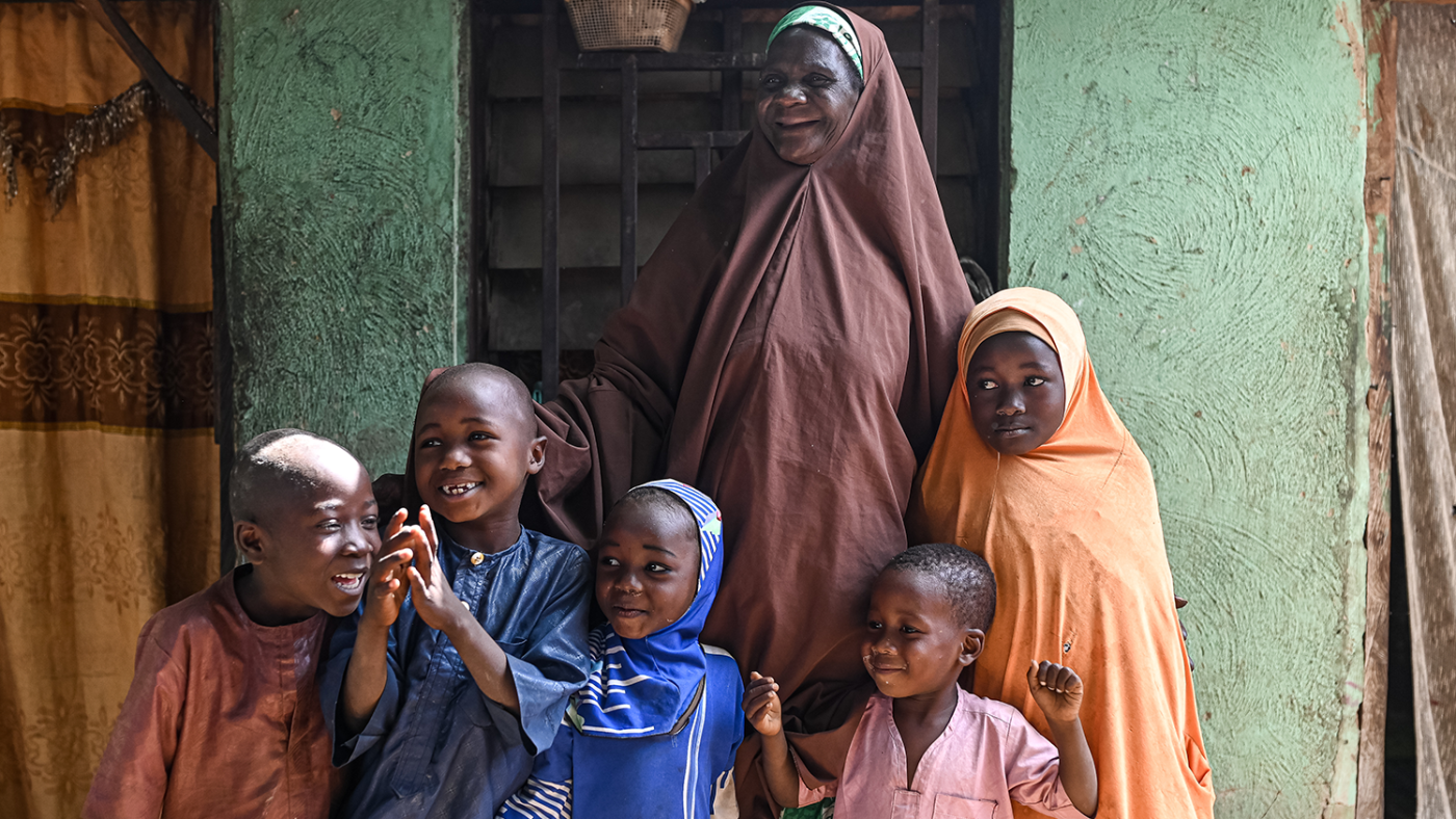 An elderly lady smiles, standing behind five young children.