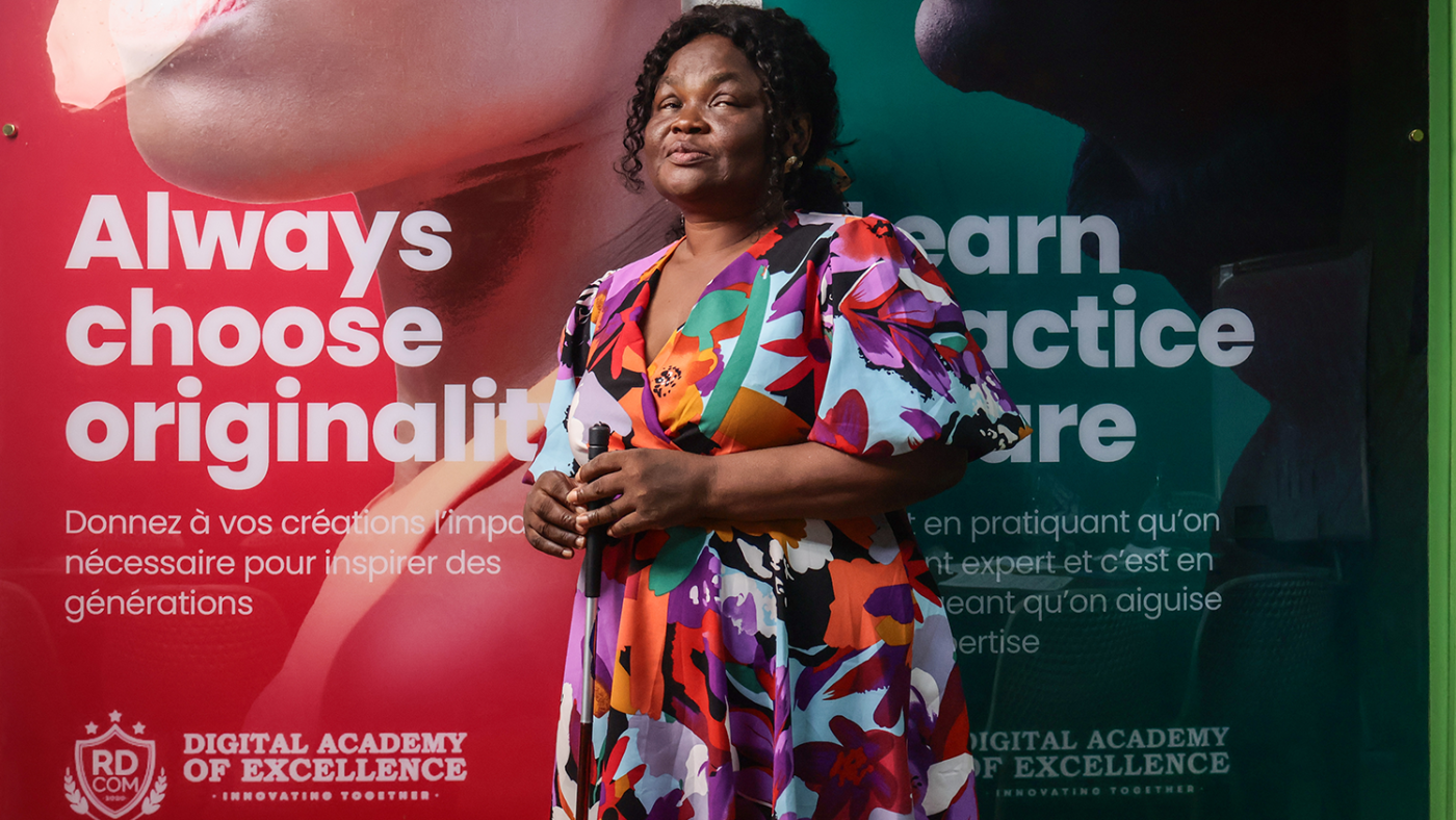 A woman wearing a colourful dress and holding a white cane stands in front of two banners.