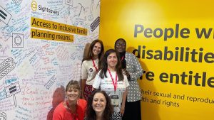 Five women pose for a group photo at a Sightsavers exhibition stand. Behind them is a wall with handwritten messages and the words 'Access to inclusive family planning means...'.