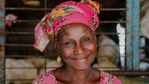 A woman wearing a pink patterned headwrap and gold earrings. Behind her, there are chickens in a cage.
