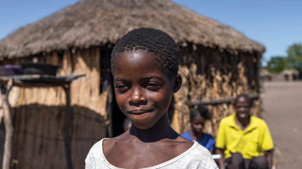 A girl stands outside a traditional grass hut. She is squinting in the bright sunshine.